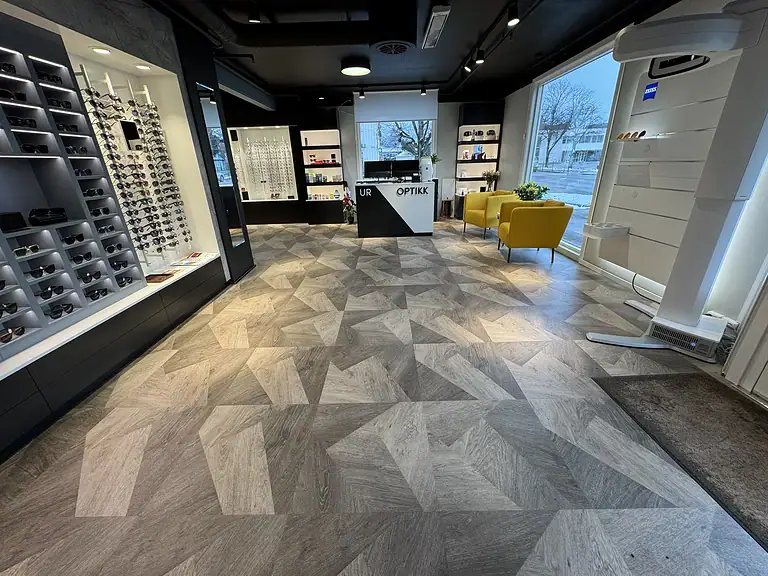 Fargerik optikkbutikk med gule komfortable stoler og stilig gulvdesign, perfekt for kjøp av briller og solbriller, moderne interiør og brede utvalg av øyepleieprodukter.