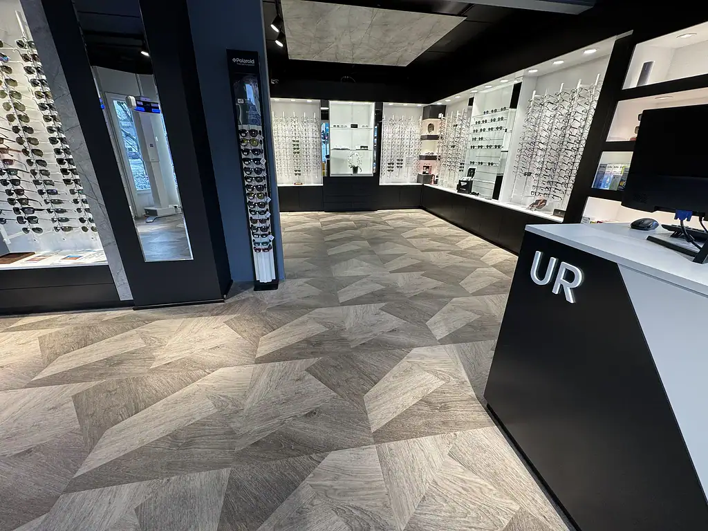 Moderne optikkbutikk innvendig med et bredt utvalg av solbriller og briller på veggene, lyst tre- og mørke skap, samt informasjonsdisplayer, fokus på kvalitet og stil.